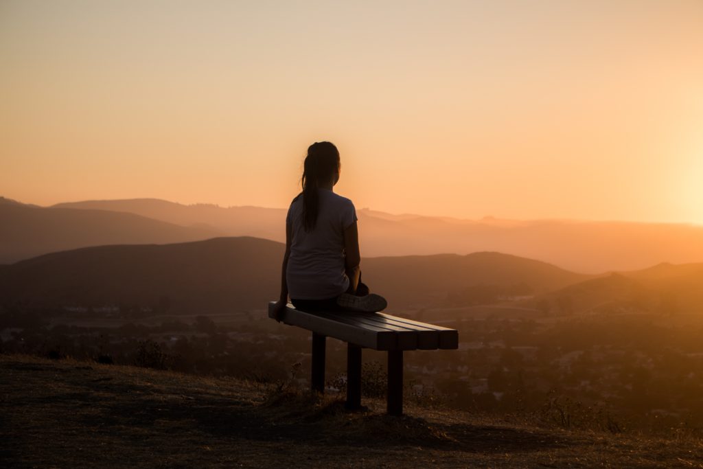 Woman spends time in nature as part of her morning routine, as she sits on a bench and looks out over mountains  at sunrise