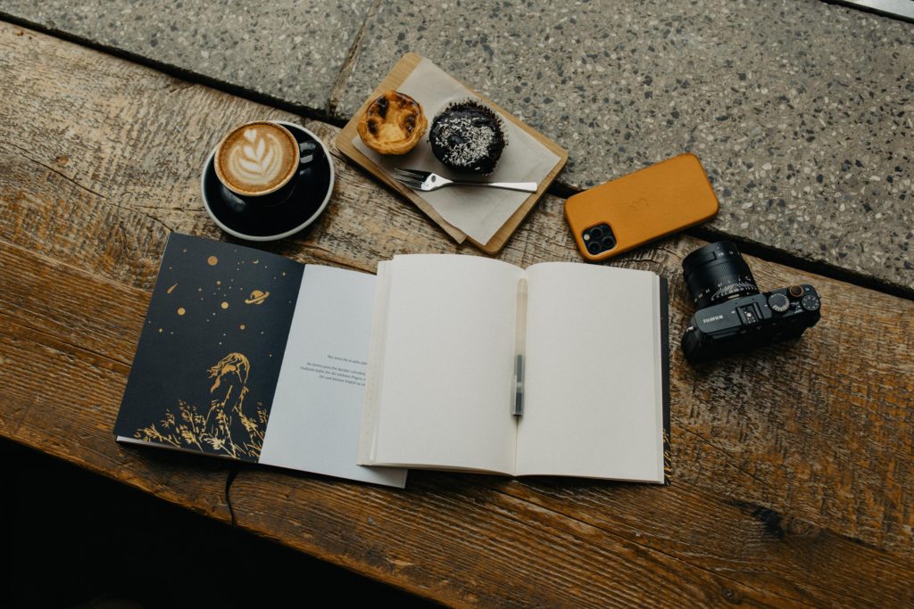 Journal with prompts open alongside photographer's camera, phone, coffee and pastries 